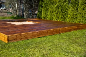 A freshly stained wooden deck features a central sandpit, surrounded by trees and bushes in a backyard.