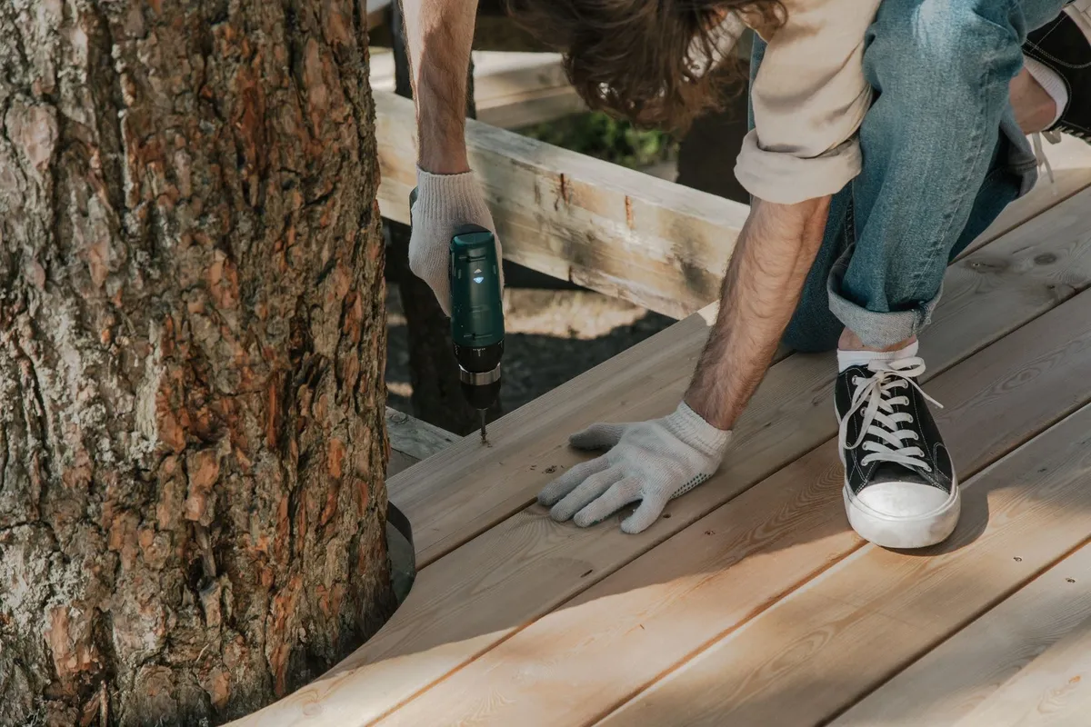 A worker drills wooden planks on a deck beside a tree, illustrating a DIY construction project.